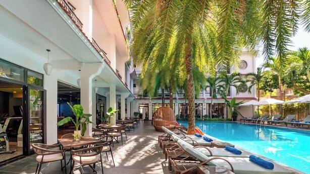 A poolside area with lounge chairs and tables, a pool, and a covered walkway with plants.