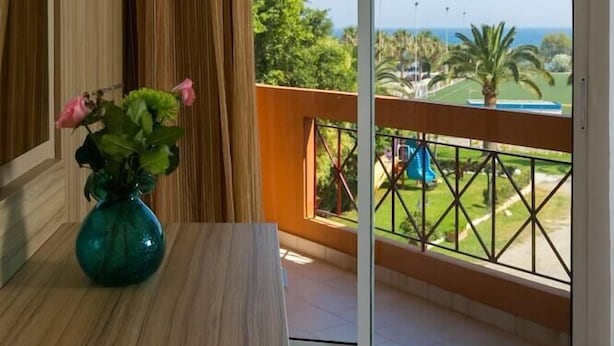 A room with a wooden table, a teal vase with flowers, and a view of a pool and greenery through a sliding glass door. - Hotel Elotia (Chania)