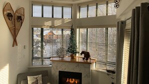 Een gezellige woonkamer met een open haard, een bank met decoratieve kussens, een kleine kerstboom en uitzicht op een besneeuwde buurt door grote ramen met jaloezieën.