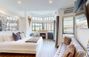 A modern bedroom with a large bed, a sofa, a fireplace, and a view of the outdoors.