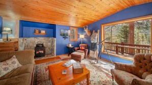 A cozy living room with a stone fireplace, wooden ceiling, and large windows overlooking a forest.
