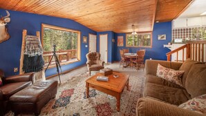 A cozy living room with a wooden ceiling, blue walls, a large window, and a rustic decor including a deer head and a tripod.