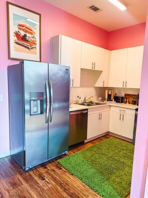 A compact kitchen with white cabinets, a stainless steel refrigerator, a dishwasher, and a green area rug.