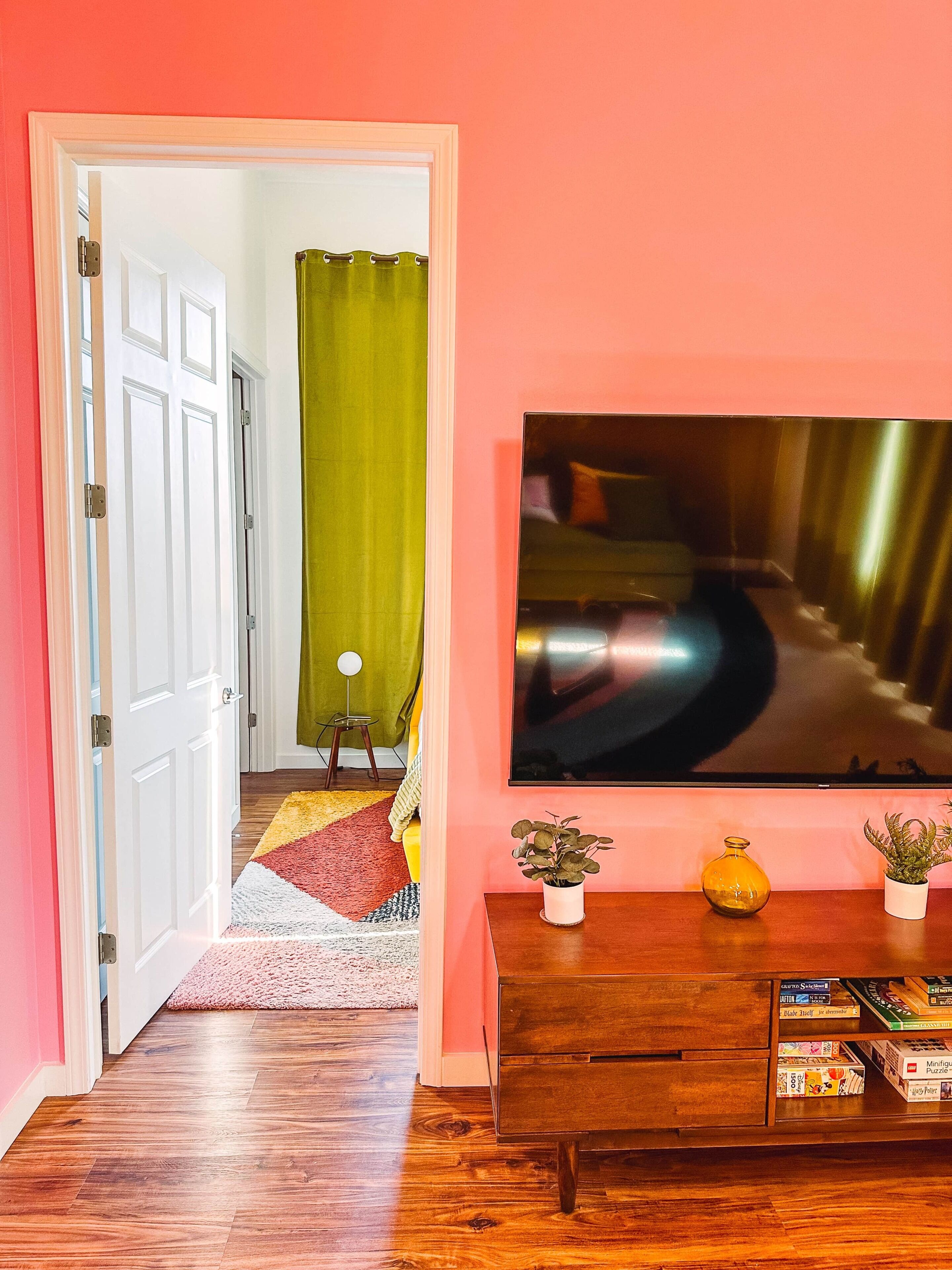 Un pasillo con piso de madera, una puerta que da a otra habitación, una alfombra colorida, una cortina verde, una pequeña mesa con una planta y un televisor montado en la pared.