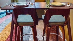 A dining area with a table set for two, two chairs, a window with curtains, and a wall with framed pictures.