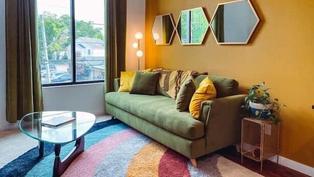 A living room with a green sofa, a glass coffee table, a colorful rug, and a large window with curtains.