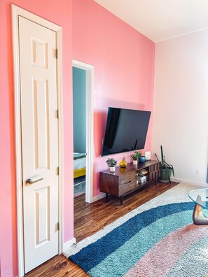 A room with a pink wall, a wooden floor, a TV on a wooden stand, a door, and a striped rug.