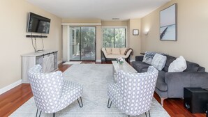 A living room with a grey sofa, two patterned armchairs, a coffee table, a flat-screen TV mounted on the wall, and a sliding glass door leading to a balcony. - Serene Courtside Villa • 2 Pools • Free Pickleball, Tennis • 5-min Walk to Beach (Hilton Head Island)