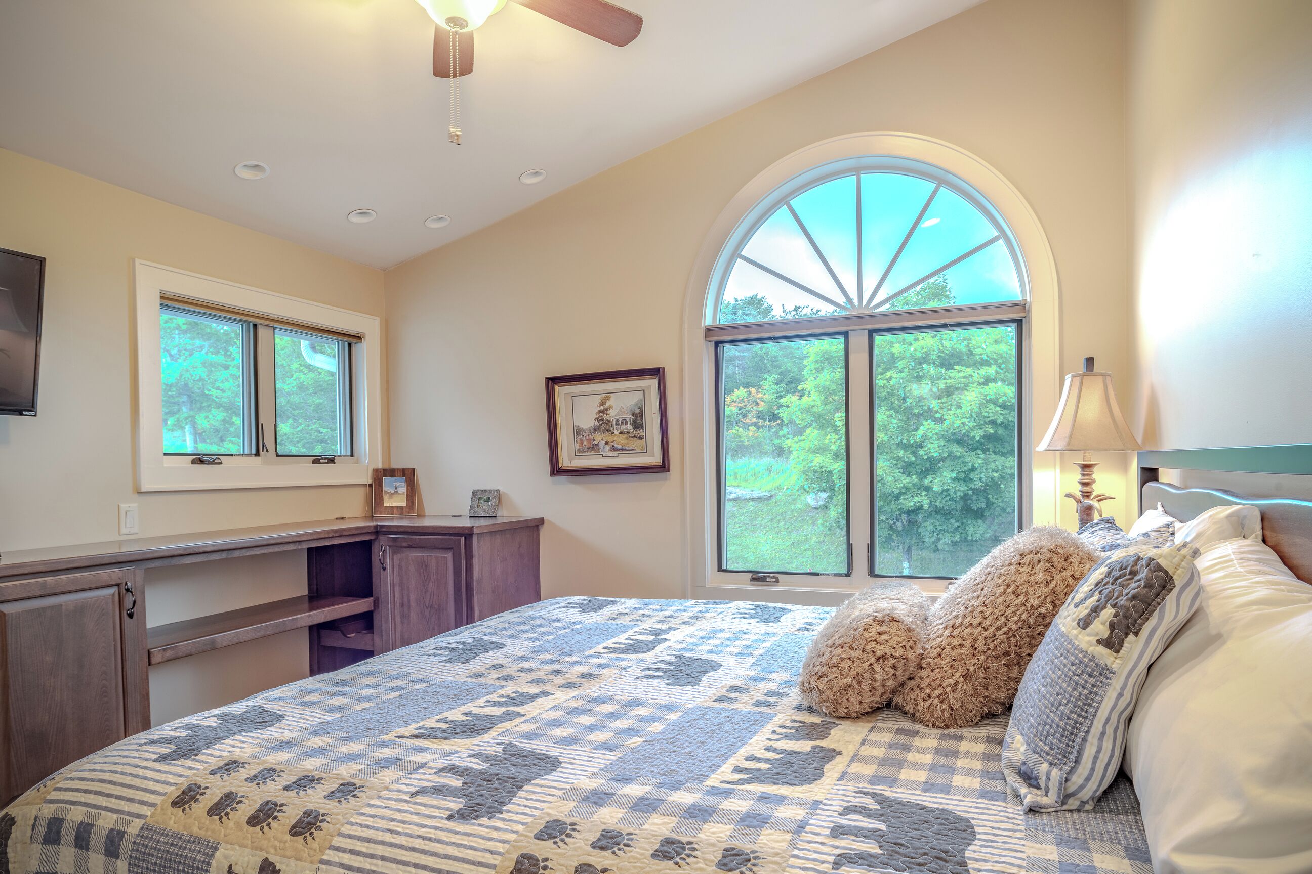 A bedroom with a large bed, a desk, a chair, a window with a view of trees, and a ceiling fan.