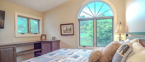 A bedroom with a large bed, a desk, a chair, a window with a view of trees, and a ceiling fan.