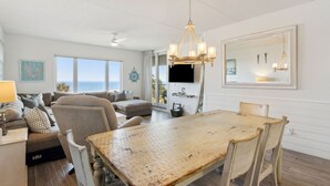 A spacious living room with a large wooden dining table, comfortable seating, a chandelier, and a view of the ocean through large windows.