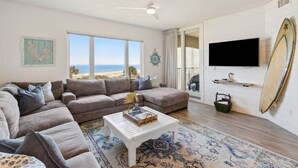 A living room with a large window, a ceiling fan, a flat-screen TV, a surfboard, and a beach view.