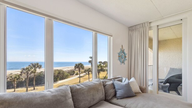 A modern living room with a large sectional sofa facing a large window overlooking a beach.