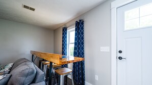 A compact living space with a wooden dining table, two bar stools, a grey sofa, and a white door.
