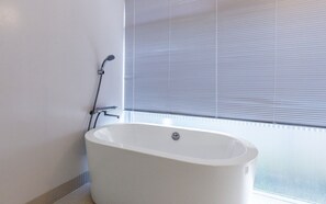 A modern bathroom with a freestanding bathtub, a showerhead, and a window with blinds. - La Maison Blanche (Yomitan)