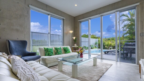 Uma sala de estar moderna com uma mesa de centro de vidro, um sofá e uma poltrona. Vê-se uma piscina e móveis de exterior através de grandes portas de correr de vidro.