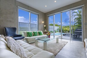 A modern living room with a glass coffee table, a sofa, and a chair. There is a view of a swimming pool and outdoor furniture through large sliding glass doors. - La Maison Blanche (Yomitan)