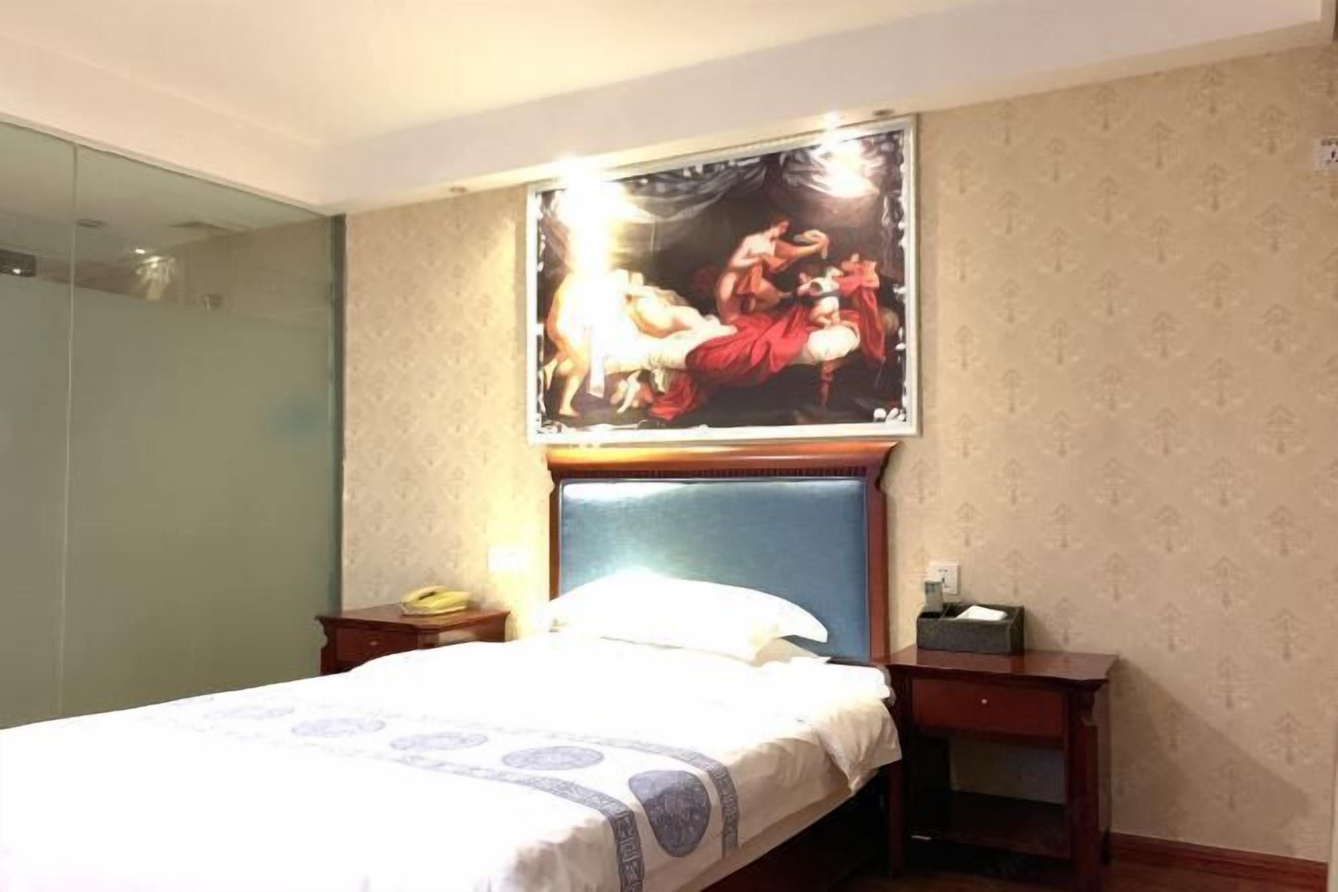 A hotel room with a bed, a nightstand, a bedside table, a glass shower enclosure, and a wall with a patterned wallpaper.