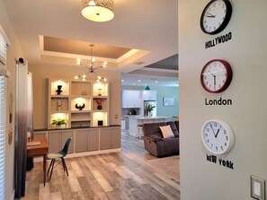 A modern living space with a kitchen, dining area, and a sofa. There are clocks labeled 'Hollywood', 'London', and 'New York' on the wall.