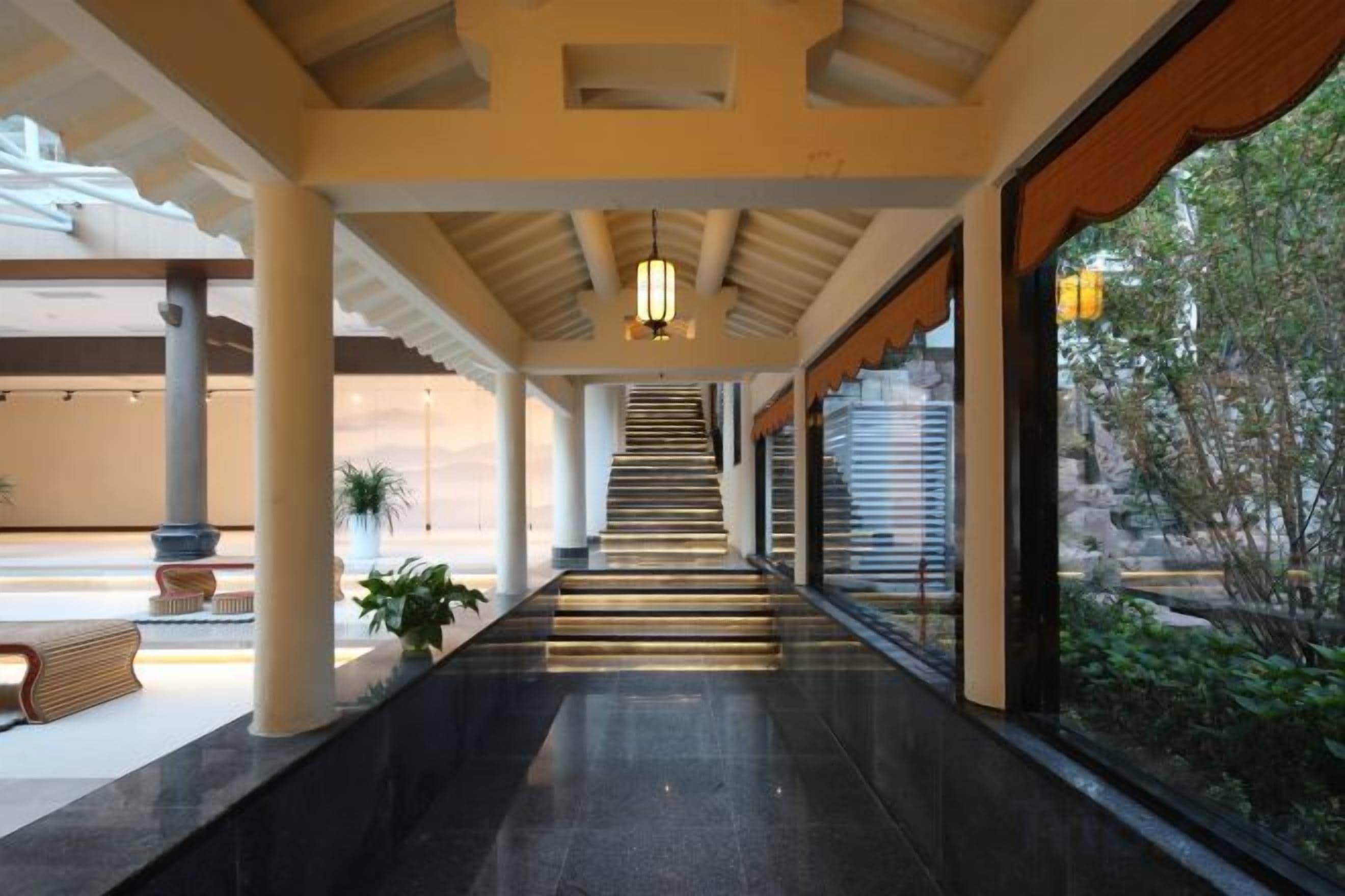 A corridor with a staircase leading to an upper level, columns, and a view of greenery outside.