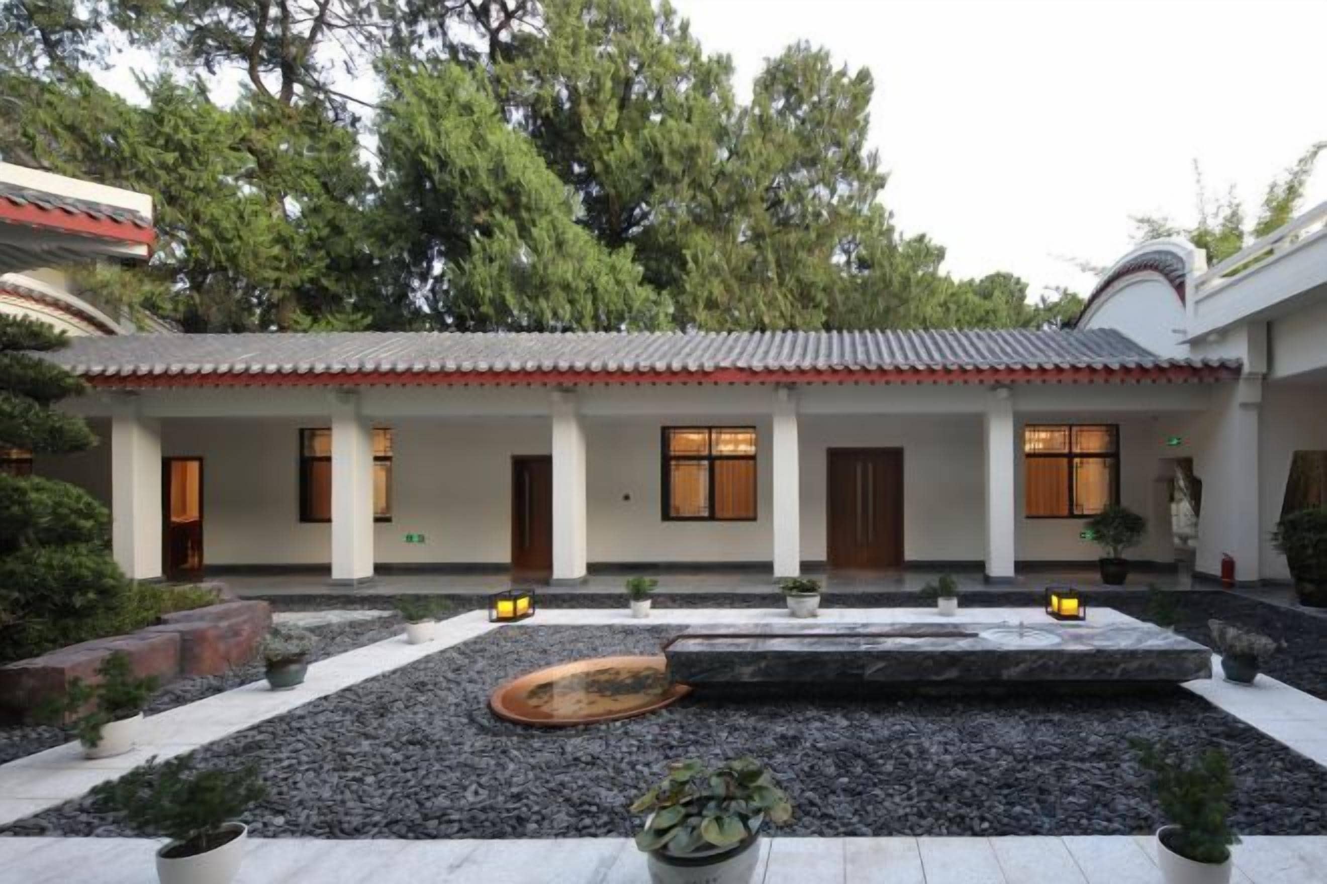 A traditional building with a tiled roof, multiple doors, and windows. There is a courtyard with gravel, plants, and a wooden bench.