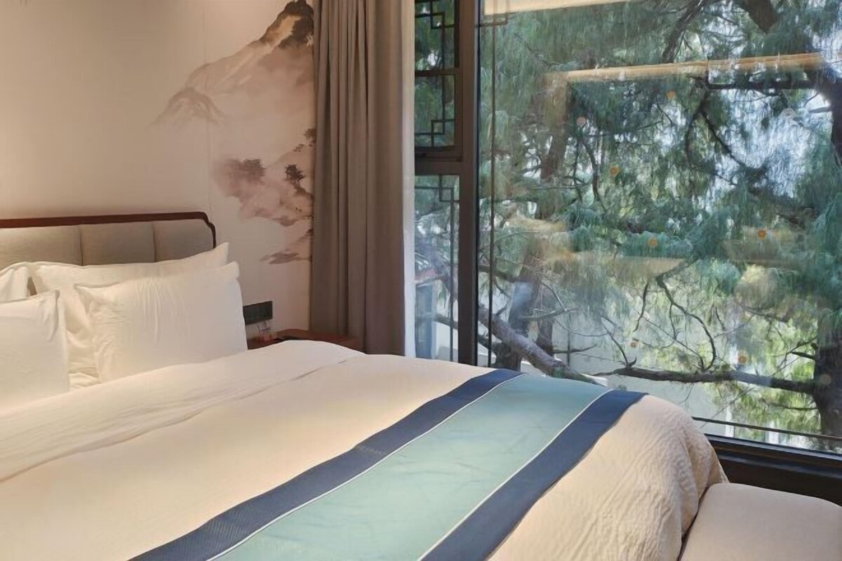 Una habitación de hotel con una cama grande, vista a árboles por una ventana y un cuadro en la pared.