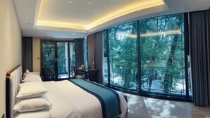 A modern hotel room with a large bed, a desk, and a view of the forest through large windows. - Qiqushan Hotel (Zitong)