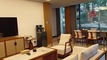 A modern hotel room with a large TV, a coffee machine, a sofa, and a dining area with chairs.