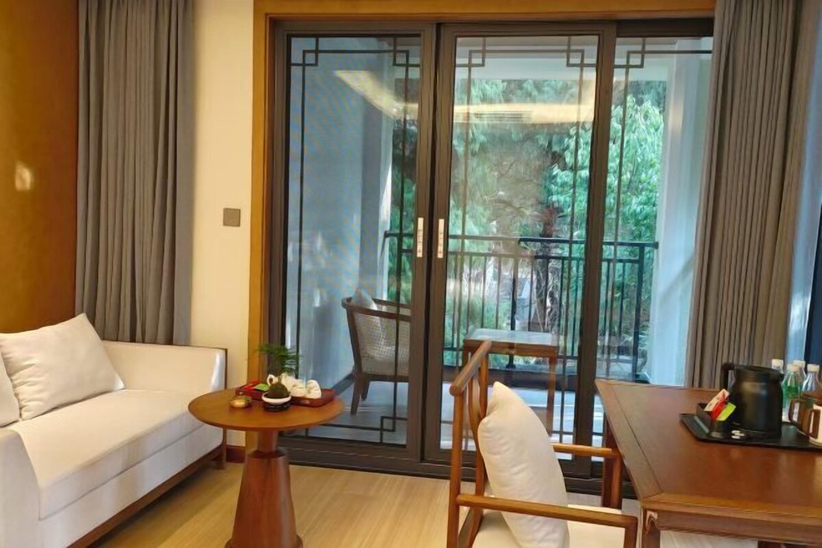 Una habitación de hotel moderna con un sofá, una mesita redonda con una planta, una silla y un balcón con vistas a zonas verdes.
