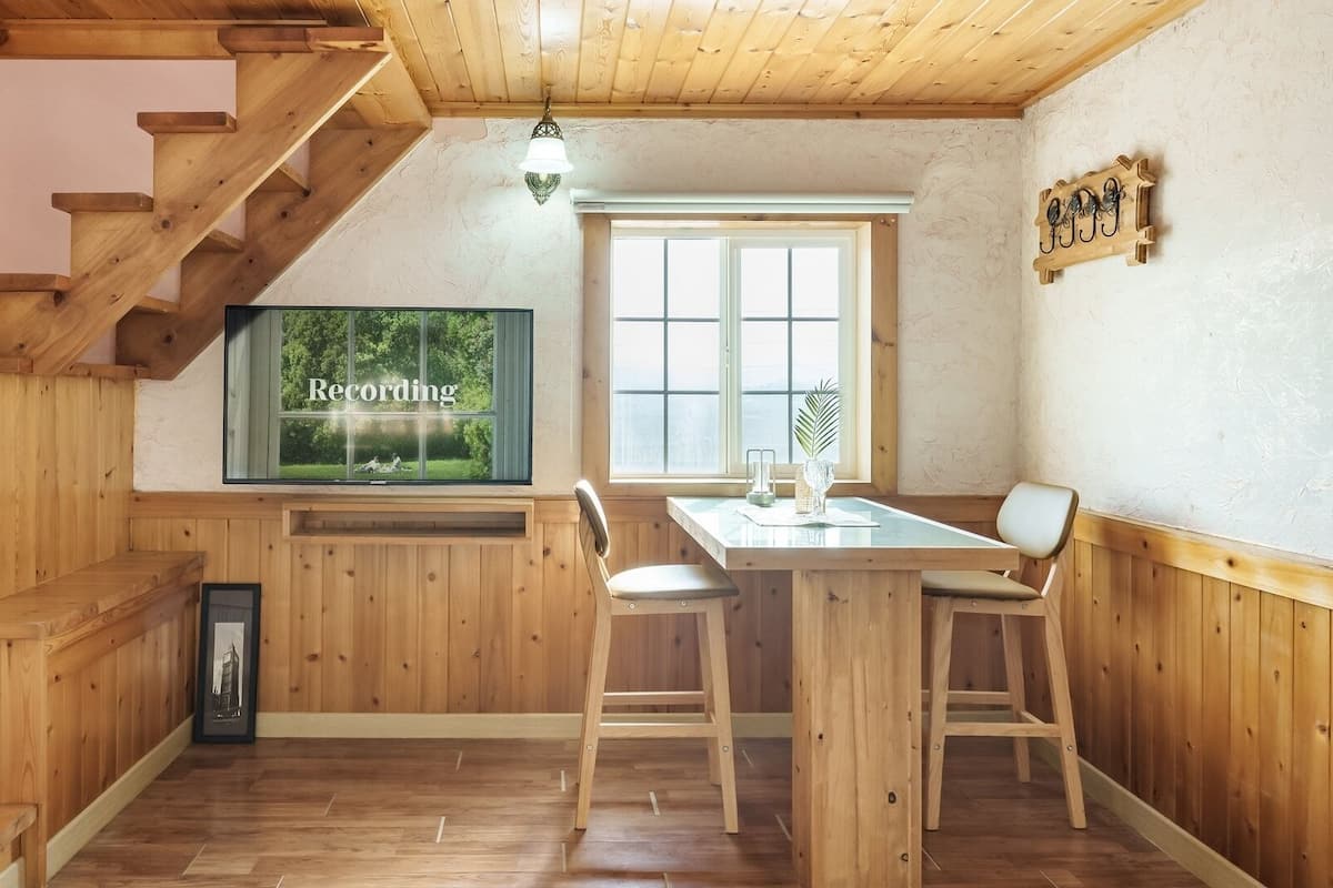 A wooden interior with a staircase, a dining area with a table and chairs, a television mounted on the wall, and a window with a view of greenery.