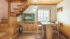 Un intérieur en bois avec un escalier, un coin repas avec une table et des chaises, un téléviseur fixé au mur et une fenêtre donnant sur un paysage verdoyant.