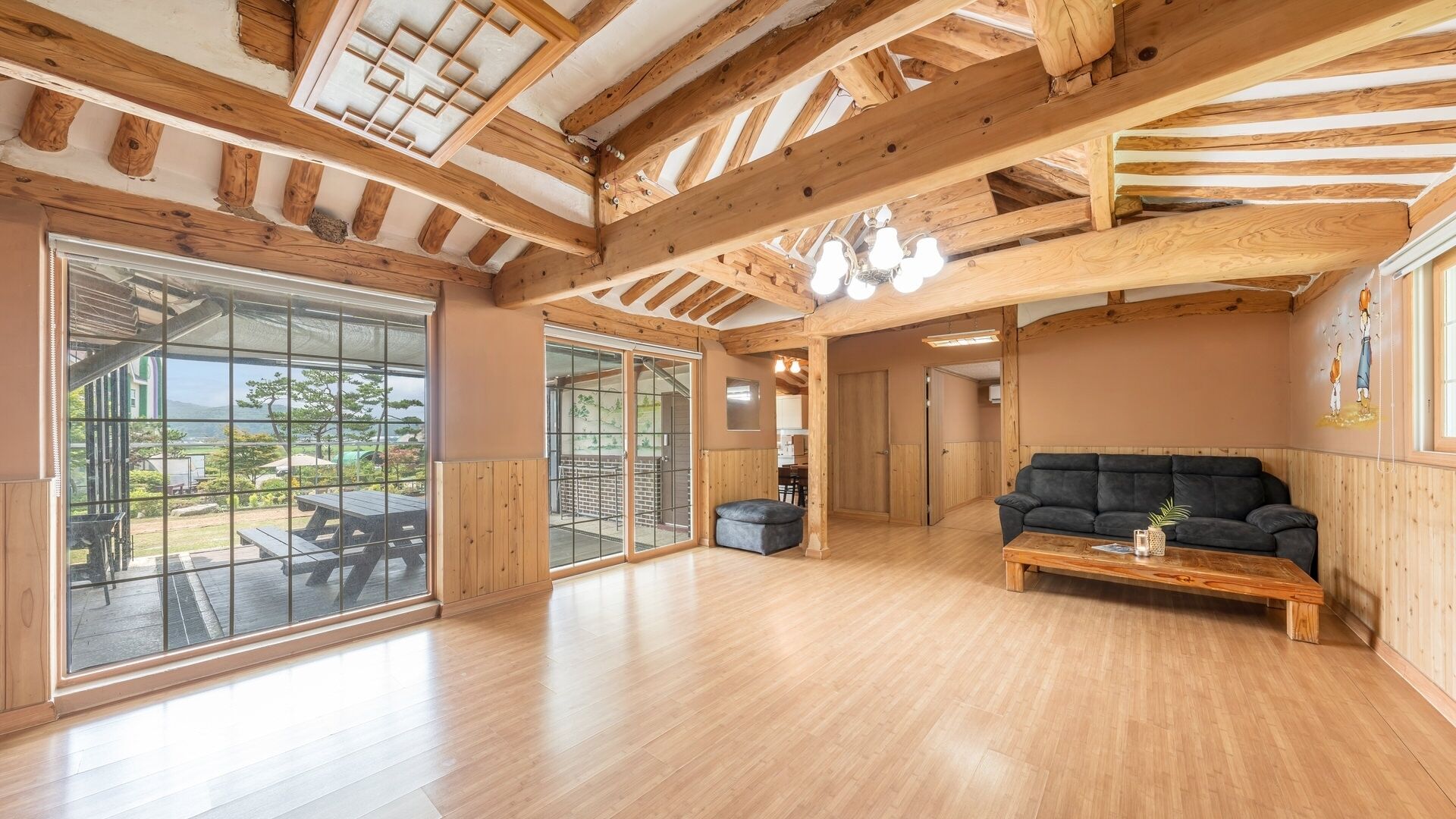 Amplia habitación con vigas de madera, un gran ventanal corredero, un sofá negro y una mesa de centro de madera.