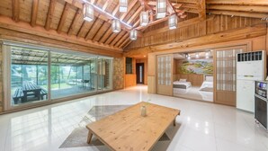 A spacious room with a wooden ceiling, large sliding glass doors leading to an outdoor patio, a wooden table, and a bed with a decorative headboard. - Ganghwa Gichayeohaeng Pension (Incheon)