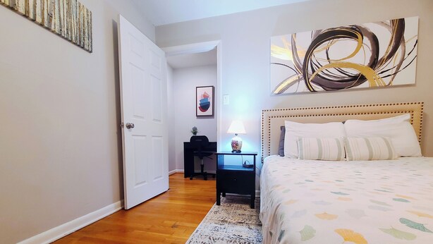 Un dormitorio con cama, mesita de noche, escritorio, silla, puerta y alfombra.