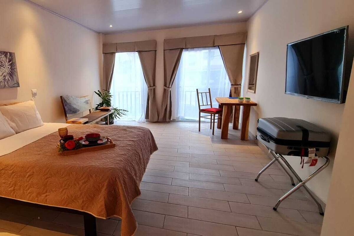 A hotel room with a bed, a TV, a desk with a chair, and a balcony with curtains.