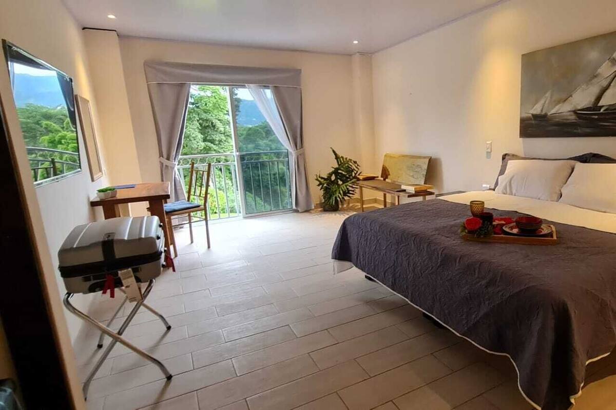 A hotel room with a bed, a desk, a chair, a balcony with a view, and a painting on the wall.