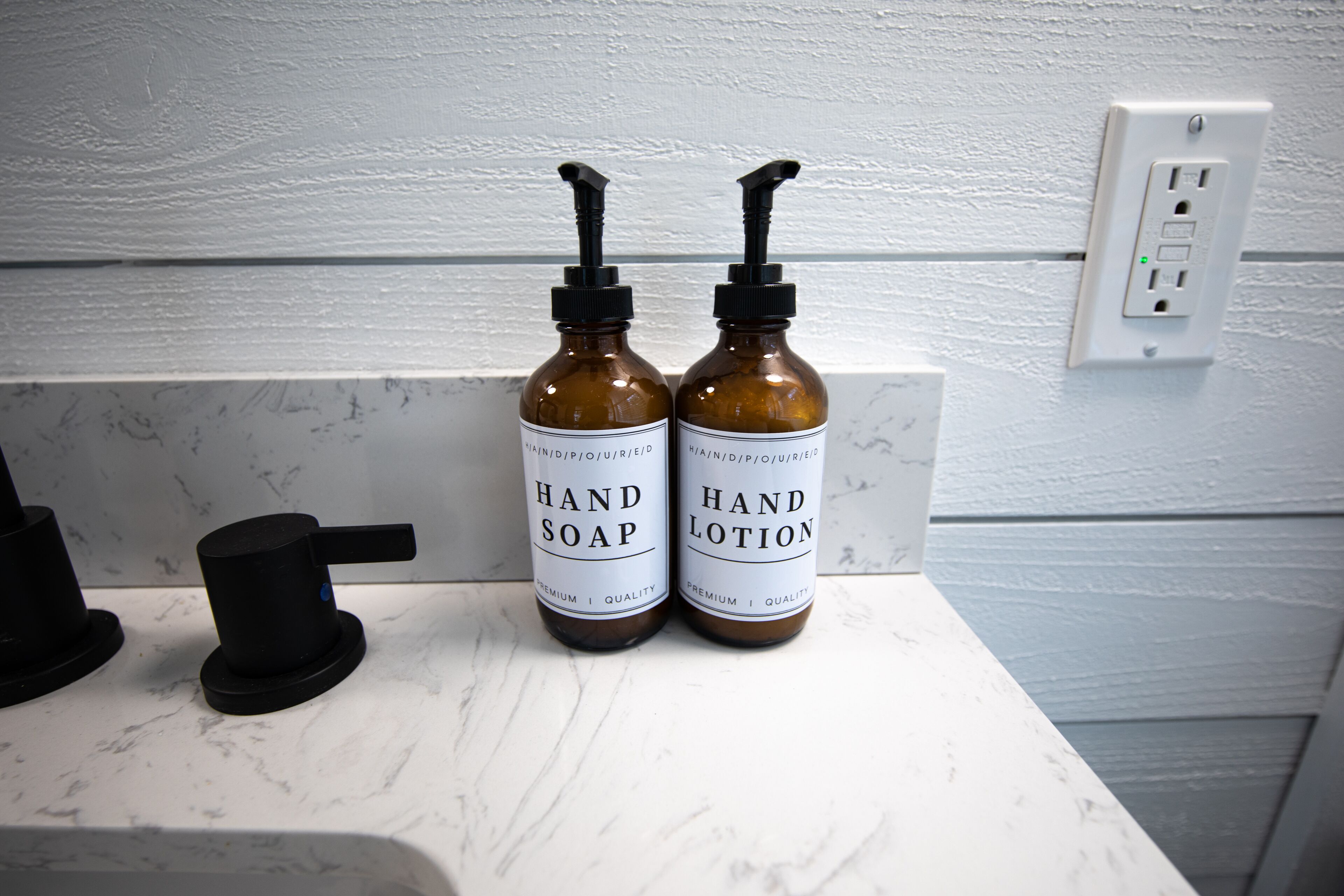 Dos botellas marrones, una con la etiqueta 'HAND SOAP' y otra con la etiqueta 'HAND LOTION', sobre una encimera de mármol blanco.