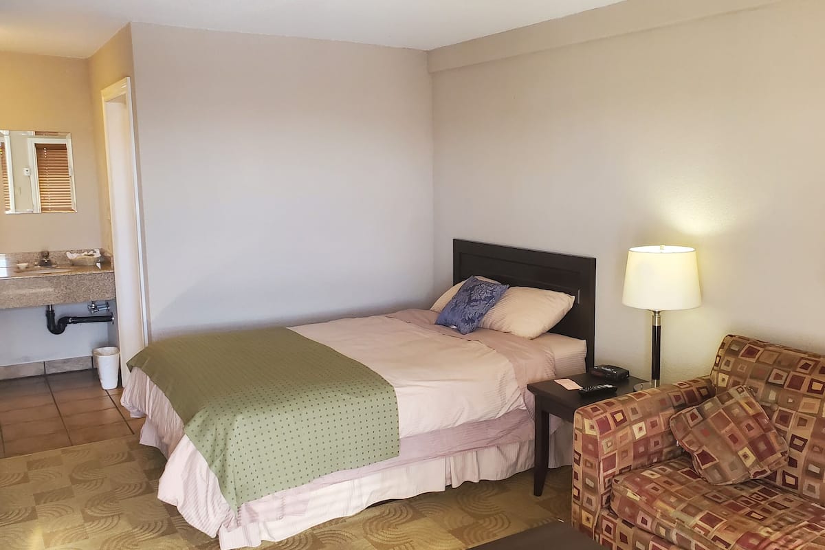A bedroom with a bed, a nightstand, a lamp, a sofa, and a bathroom visible through an open door.