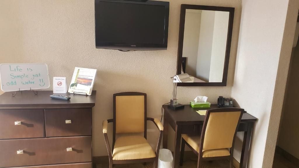 A hotel room with a desk, two chairs, a television, a mirror, and a sign with a motivational quote.