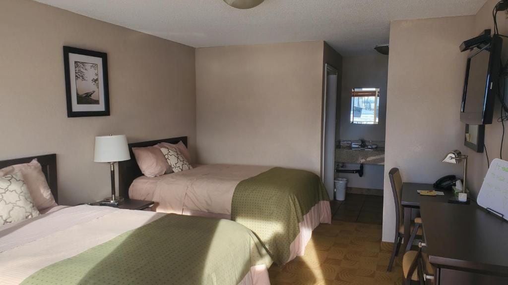 Ein Hotelzimmer mit zwei Betten, einem Schreibtisch, einem Stuhl, einer Lampe, einem gerahmten Bild an der Wand und einem durchgehenden Tür sichtbaren Badezimmer.
