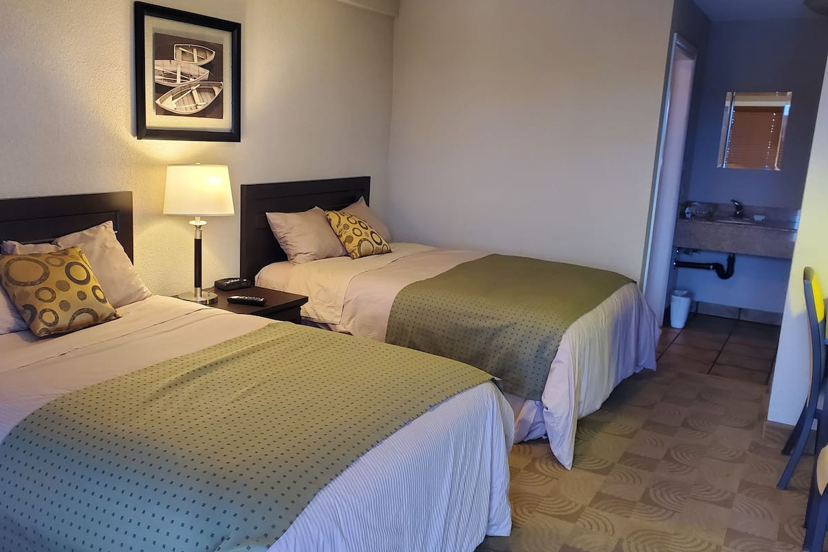 A hotel room with two beds, a nightstand with a lamp, a framed picture on the wall, and a bathroom visible through an open door.