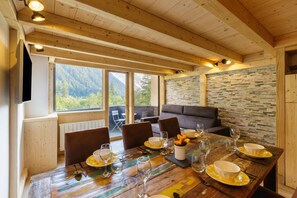 A wooden dining table set with plates, glasses, and cutlery, with a stone wall and a view of mountains in the background.