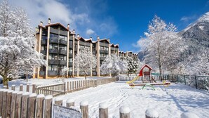 A snow-covered outdoor area with a wooden fence, a playground, and a building with multiple balconies. - Grand Roc A216 150m from Slopes, Mont-Blanc View & Parking, Argentière (Chamonix), France (Argentière (Chamonix))