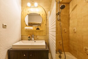 A bathroom with a white sink, wooden vanity, a mirror with lights, a shower with a handheld showerhead, and floral patterned tiles. - Appartement Central Des Houches - 15, Les Houches (Chamonix), France (Les Houches (Chamonix))