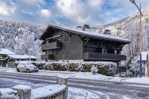 Un edificio de dos pisos con balcón, rodeado de árboles y montañas cubiertas de nieve.