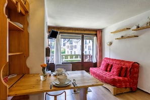 A cozy room with a wooden dining table, a red sofa, and a window with a view of buildings. - Appartement Central Des Houches - 15, Les Houches (Chamonix), France (Les Houches (Chamonix))