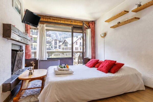 A hotel room with a bed, a small table with a decorative item, a fireplace, and a view of snow-covered buildings.