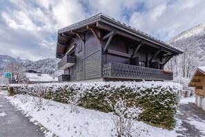 A two-story wooden chalet with a balcony, surrounded by snow-covered trees and mountains.