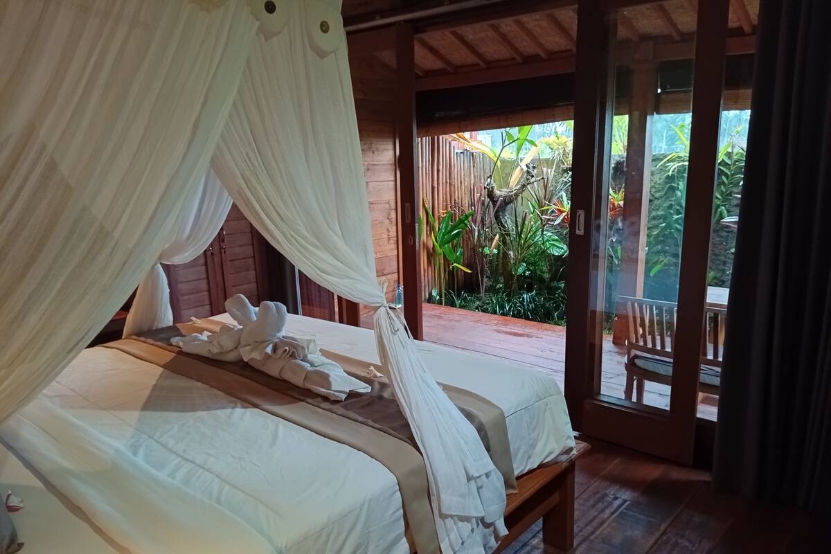 Quarto com duas camas, piso de madeira e vista para um jardim através de uma porta aberta.