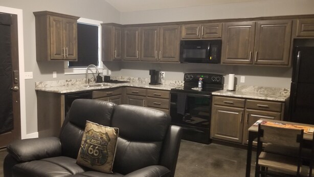A kitchen with dark wooden cabinets, a black microwave, and a black stove. There is a black leather chair with a pillow. - New 1 bedroom cottage right off Route 66. (Galena)